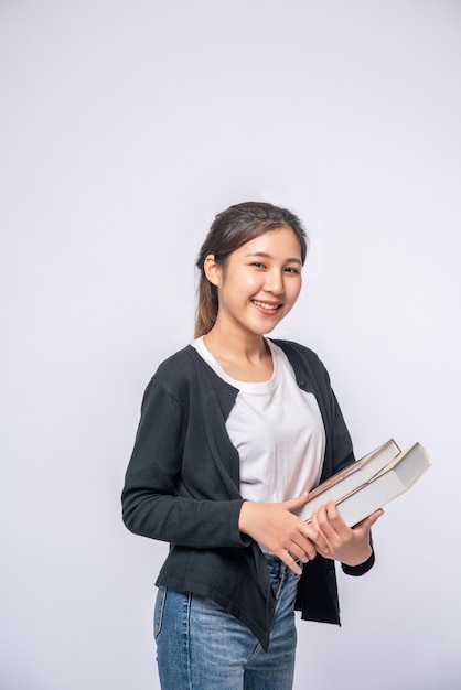 Mujer joven sonriendo felizmente con una camisa negra y jeans, sosteniendo un libro y sonriendo felizmente.