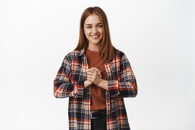 Mujer joven sonriendo con las manos apretadas cerca del pecho esperanzada, pidiendo algo, esperando, rogando tener una oportunidad, de pie contra el fondo blanco