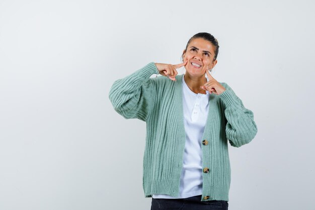 Mujer joven sonriendo y poniendo los dedos índices en las mejillas con camisa blanca y chaqueta de punto verde menta y mirando alegre