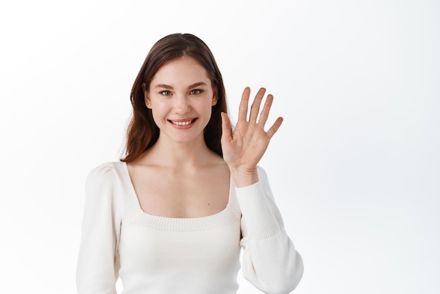 Mujer joven sonriendo y saludando saludando con la mano para saludar a los amigos decir hola y dar la bienvenida adiós gesto de pie contra el fondo blanco.