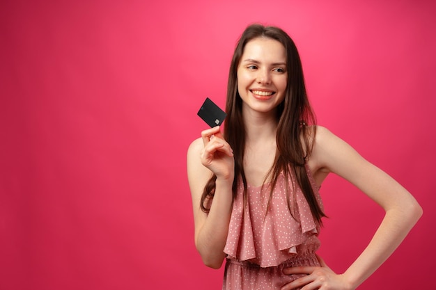 Mujer joven sonriente atractiva que sostiene la tarjeta de crédito negra contra el fondo rosado del estudio