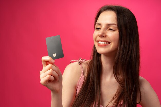 Mujer joven sonriente atractiva que sostiene la tarjeta de crédito negra contra el fondo rosado del estudio