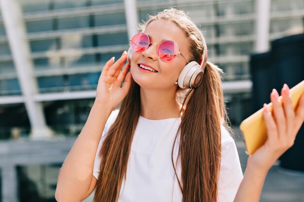 Mujer joven sonriente y bailando sosteniendo un teléfono inteligente y escuchando música en auriculares