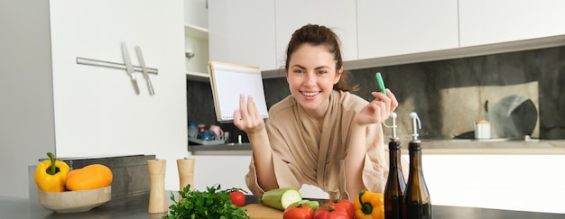 Foto gratuita mujer joven sonriente en bata de baño cocinando en la cocina parada cerca de verduras con cuaderno