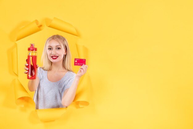 Mujer joven sonriente con botella roja y tarjeta de crédito sobre fondo amarillo deporte atleta gimnasio dinero