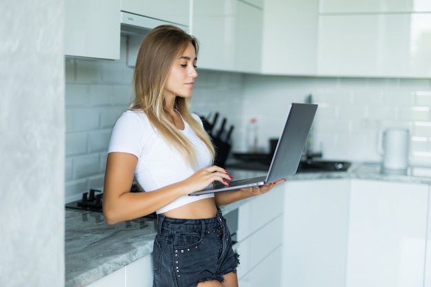 Mujer joven sonriente en la cocina que mira su computadora portátil