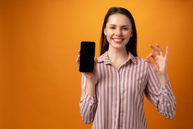 Mujer joven sonriente feliz que le muestra la pantalla negra del teléfono inteligente con espacio de copia