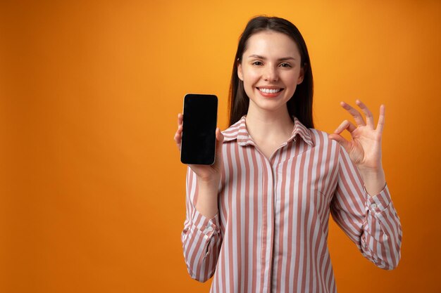 Mujer joven sonriente feliz que le muestra la pantalla negra del teléfono inteligente con espacio de copia