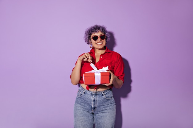 Mujer joven sonriente con gafas de sol con borde rojo, aretes redondos de oro y camiseta roja se regocija de su regalo en lila.