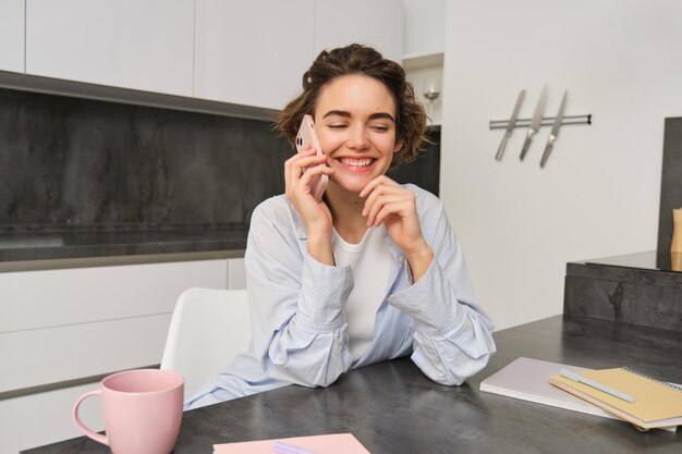 Mujer joven sonriente habla por teléfono móvil llama a alguien de casa se sienta en la cocina y tiene conversati