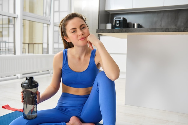 Foto gratuita una mujer joven sonriente haciendo deportes en su casa mirando a la cámara y sonriendo bebiendo agua después