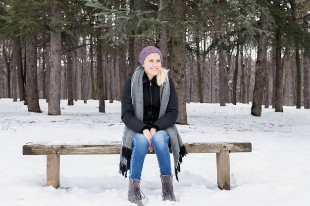 Mujer joven sonriente hermosa que se sienta en banco de madera en bosque del invierno