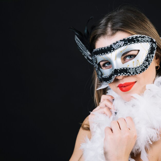 Mujer joven sonriente en la máscara del carnaval que sostiene la pluma de la boa en fondo negro
