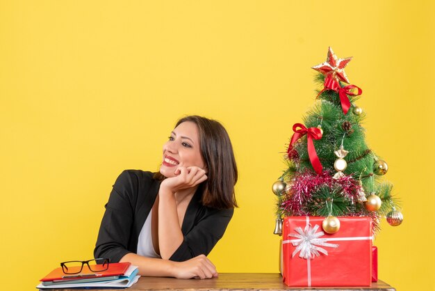 Mujer joven sonriente mirando algo sentado en una mesa cerca del árbol de Navidad decorado en la oficina en amarillo
