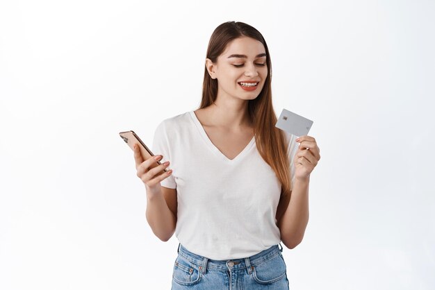 Mujer joven sonriente mirando el número de la tarjeta de crédito mientras paga en la aplicación de transferencia de dinero del teléfono móvil comprando en línea en el teléfono inteligente usando la aplicación bancaria de pie sobre fondo blanco