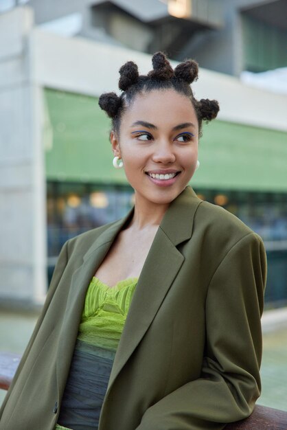 La mujer joven sonriente de moda con moños vestida con chaqueta verde usa maquillaje vívido mira hacia otro lado felizmente disfruta del tiempo libre en la ciudad posa al aire libre contra el fondo borroso se siente feliz.