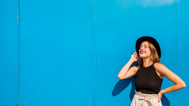 Mujer joven sonriente moderna que presenta delante de la pared azul