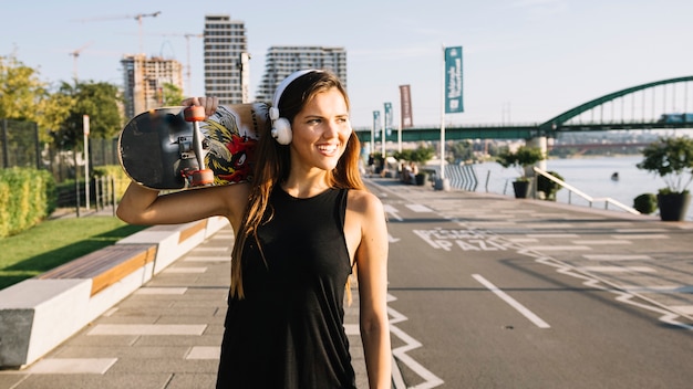 Mujer joven sonriente con el monopatín que escucha la música en el auricular