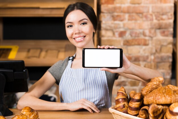 Mujer joven sonriente en el mostrador de la panadería que muestra su teléfono móvil