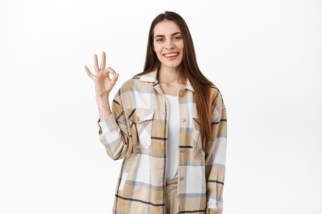 La mujer joven sonriente muestra el signo de estar bien y asiente con la cabeza en señal de aprobación, se ve complacida, satisfecha con la buena calidad, elogia el buen trabajo, excelente elección, se para sobre la pared blanca