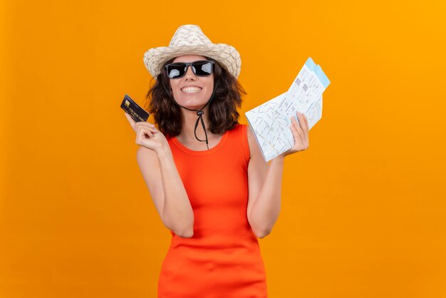 Una mujer joven sonriente con el pelo corto en una camisa naranja con sombrero para el sol y gafas de sol con mapa y tarjeta de crédito