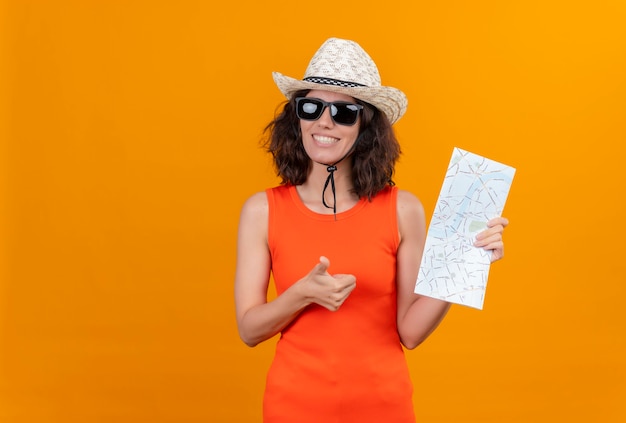 Una mujer joven sonriente con el pelo corto en una camisa naranja con sombrero para el sol y gafas de sol sosteniendo el mapa mirando felizmente mostrando los pulgares para arriba