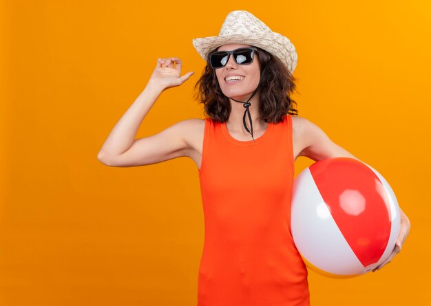 Una mujer joven sonriente con el pelo corto en una camisa naranja con sombrero para el sol y gafas de sol sosteniendo una pelota inflable mostrando adiós con la mano