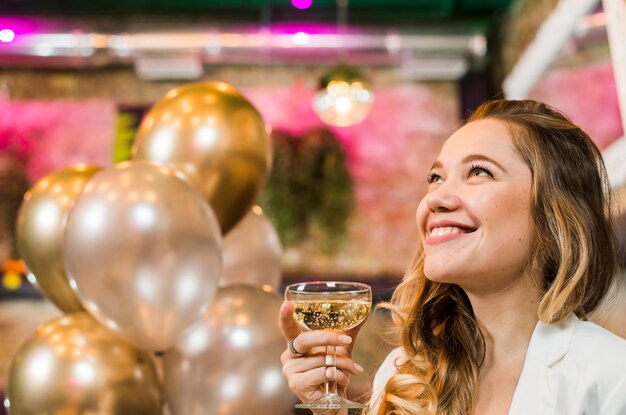 Mujer joven sonriente pensativa que sostiene el vidrio del whisky en barra