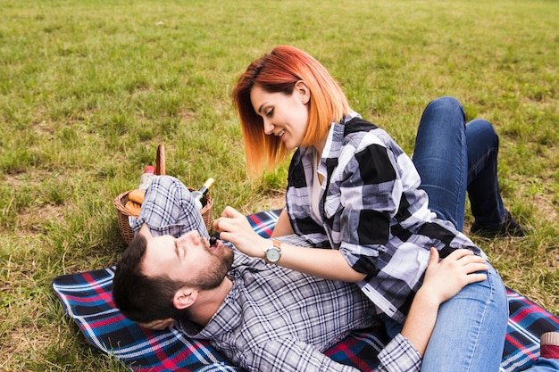 Mujer joven sonriente que alimenta la cereza a su novio que miente en la manta sobre hierba verde