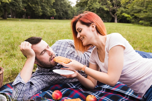 Mujer joven sonriente que alimenta la pasta de hojaldre a su novio en la comida campestre