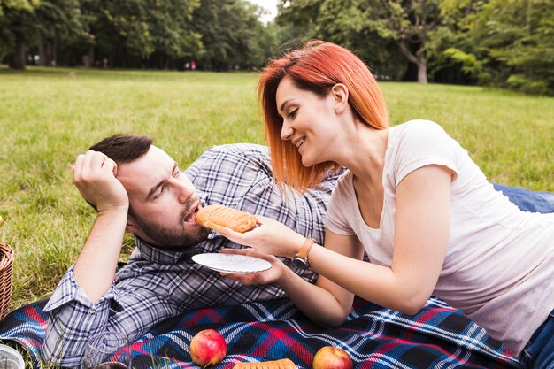 Mujer joven sonriente que alimenta la pasta de hojaldre a su novio en la comida campestre