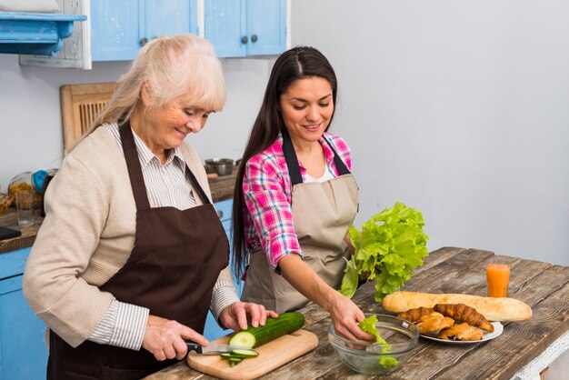 Mujer joven sonriente que ayuda a su madre mayor para preparar la ensalada en la cocina