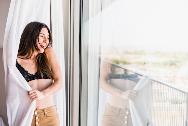 Mujer joven sonriente que se coloca en cortina cerca de la ventana