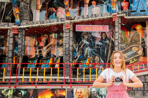 Mujer joven sonriente que se coloca delante de la diversión de la casa encantada paseo que sostiene la cámara