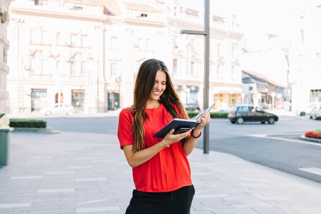 Mujer joven sonriente que se coloca en el diario de la lectura de la acera