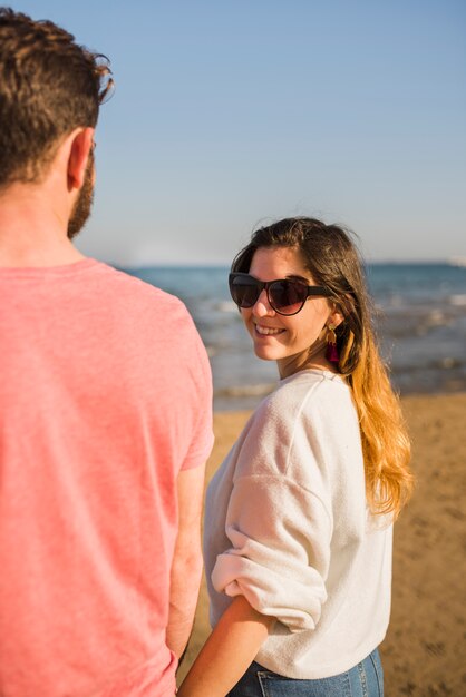 Mujer joven sonriente que se coloca con sus gafas de sol que llevan del novio que miran sobre hombro en la playa