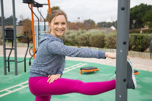 Mujer joven sonriente que estira la pierna en el campo de deportes