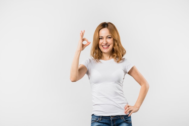 Mujer joven sonriente que hace gesto aceptable contra el contexto blanco