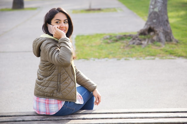 Mujer joven sonriente que llama en el teléfono y que da vuelta detrás en parque