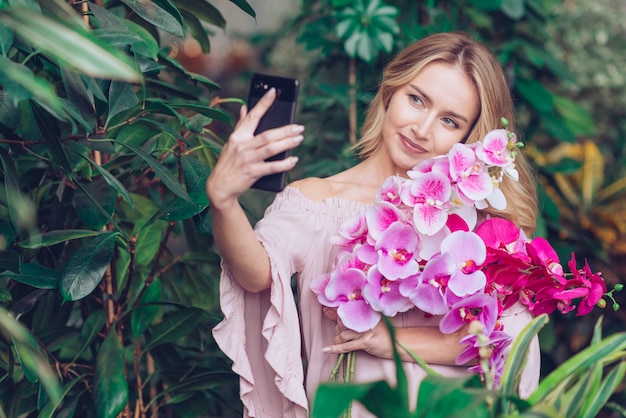Mujer joven sonriente que lleva a cabo las ramas hermosas de la orquídea en la mano que toma el selfie en el teléfono elegante