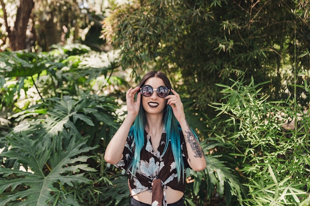 Mujer joven sonriente que lleva las gafas de sol que se colocan entre árboles