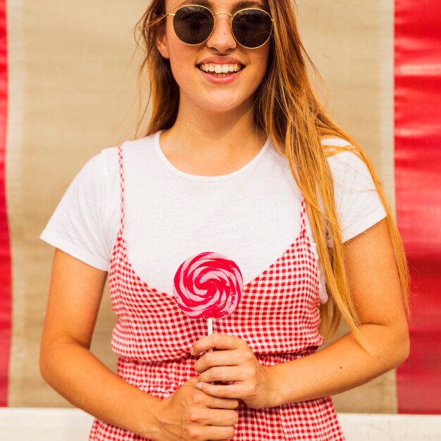 Mujer joven sonriente que lleva las gafas de sol que sostienen la piruleta disponible