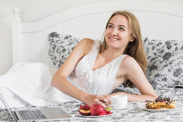 Mujer joven sonriente que miente en cama que goza del desayuno de la mañana