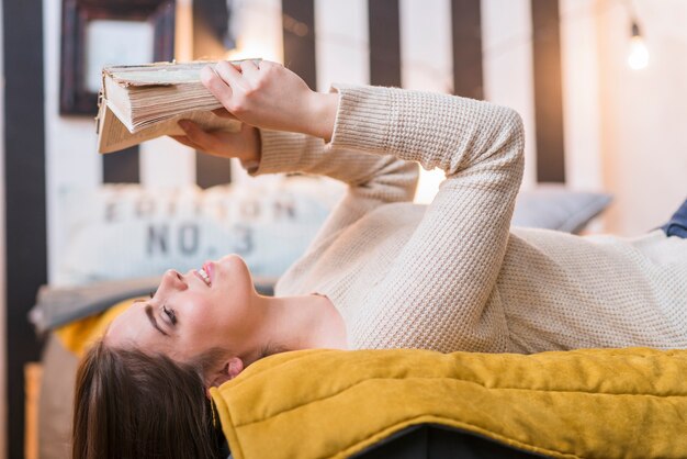 Mujer joven sonriente que miente en cama que lee el libro