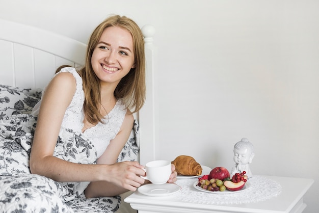 Mujer joven sonriente que miente en la cama que sostiene la taza de café con el desayuno en la tabla lateral