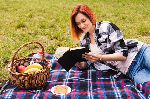 Mujer joven sonriente que miente en el libro de lectura de la manta en la comida campestre