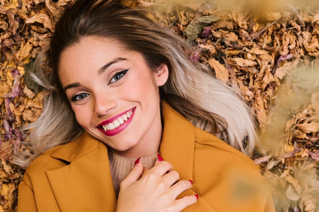 Mujer joven sonriente que mira la cámara que miente en las hojas secas