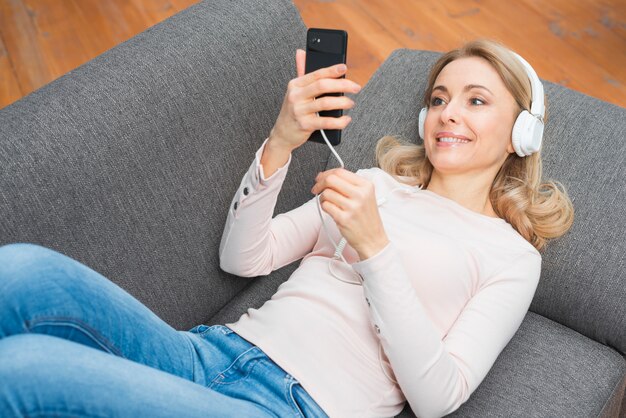 Mujer joven sonriente que mira música que escucha del teléfono móvil en el auricular