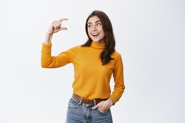 Mujer joven sonriente que muestra una mano de tamaño pequeño y mirando una cosita con expresión de cara feliz y satisfecha, de pie en ropa casual en blanco