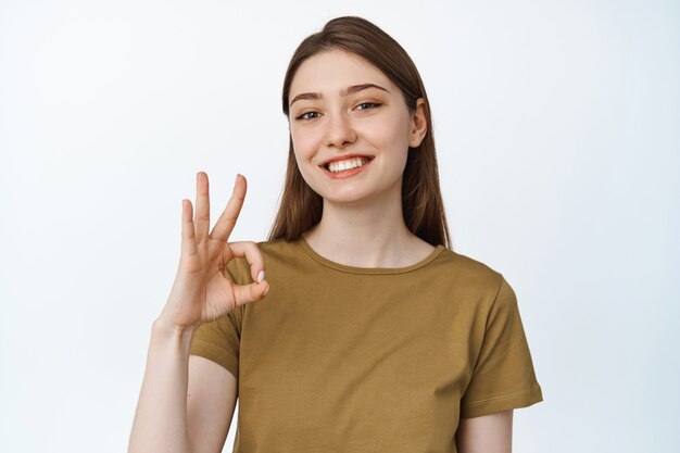 Mujer joven sonriente que muestra el signo de ok, ok y luciendo satisfecho, recomendando algo, felicitando la calidad, de pie satisfecho en blanco